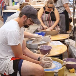 A group of students at ceramics wheels, one in the foreground is throwing a small bowl